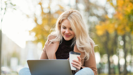 Virtual communication. Happy woman. Distance connection. Smiling lady sitting in autumn park with cup of coffee typing laptop blurred nature view.