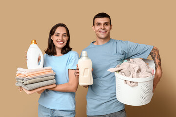 Young couple with laundry basket and bottles of detergent on beige background