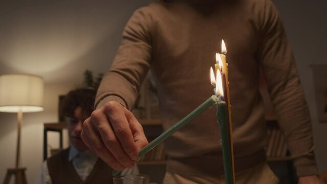 Close-up shot of hand of unrecognizable young man holding shammash and lighting up all candles on hanukkiah on final day of Hanukkah holiday, and young boy in kippah sitting in background
