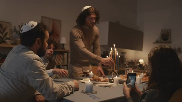 Medium shot of widowed middle-aged Jewish man in kippah celebrating Hanukkah at home with his children, eldest son lighting up candles on menorah and young daughter filming on smartphone
