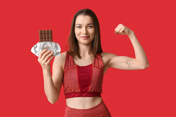 Young woman holding chocolate bar and showing muscles on red background