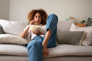 Young multiracial woman watching movie at home eating popcorn, sitting on the couch with surprise...