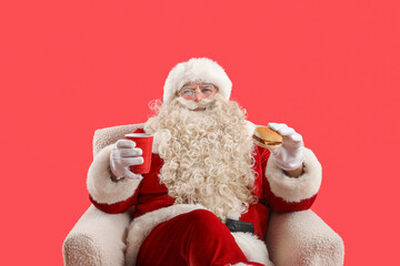 Santa Claus with cup of tea and tasty burger in armchair on red background