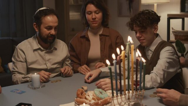 Medium shot of teenage Jewish boy wit curly hair, in kippah playing dreidel with family during Hanukkah feast, spinning, getting Hebrew letter Hei, meaning half, taking gelt coins, and all laughing