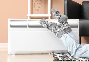 Young woman in warm socks near electric heater at home. Concept of heating season