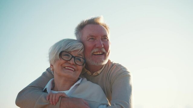 Happy laughing senior man hugging woman on sea beach at sunset in evening. Elderly couple family on date cuddling embracing talking. Lifelong relationships happy marriage, love enjoy leisure concept.