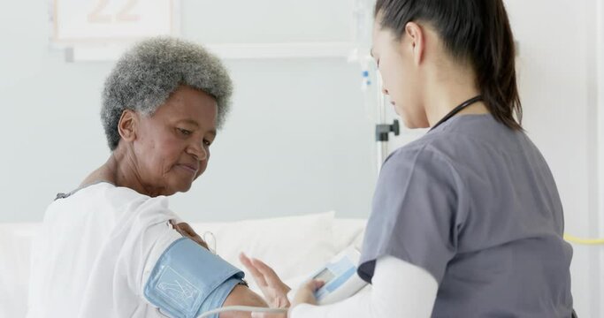 Diverse female doctor testing blood pressure of senior female patient in hospital room, slow motion