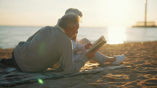 Senior gray haired man woman reading book together sitting on sandy sea beach at sunset in evening. Happy marriage, old family spending enjoy time. Love couple relationship, leisure, resting concept.