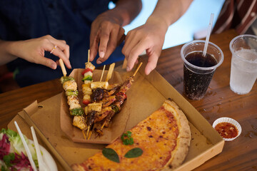 Boxes with meat skewers and pizza on table in front of friends, view from above