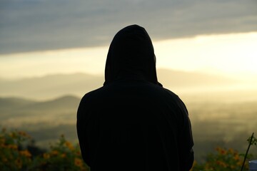 A backlit photograph of a man standing in a hoodie looking at the evening mountain view.
