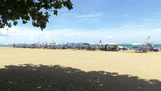 Beachfront footage taken from under the shades of trees capturing a line of beach canopies with colorful banners overlooking the sea and mountains.
