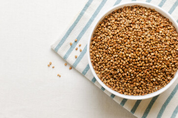 Organic dry buckwheat groats in a white bowl on a white striped napkin on a light background. Vegetarian and vegan food. Concept of healthy eating. Horizontal orientation. Top view. Copy space.