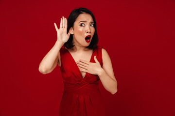 Amazed asian woman trying to hear something while keeping hand at her ear isolated over red wall