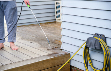 Man cleaning wooden deck with hand-held pressure sprayer. Garden hose in the foreground.