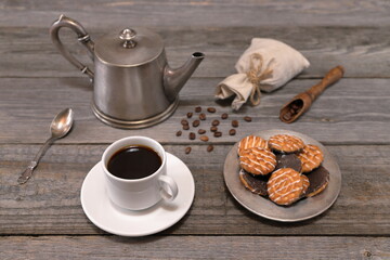 A still life with white cup of coffee, plate with biscuits, coffee pot, wooden scoop with coffee beans and scattering of coffee beans in front of small burlap bag there is on gray wooden background. 