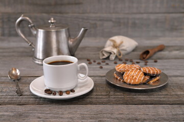 A still life with white cup of coffee, plate with biscuits, coffee pot, wooden scoop with coffee beans and scattering of coffee beans in front of small burlap bag there is on gray wooden background. 