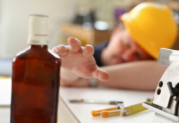 Arm of drunken worker in yellow helmet hold liquor bottle sleeping at table closeup. Manual job...