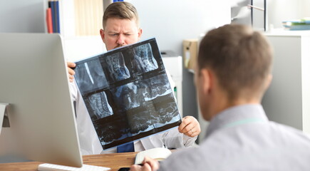 Portrait of serious doctor looking carefully at spine radiograph in different projections. Ill man...