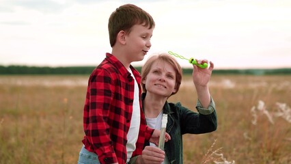 Cheerful boy in checkered shirt blows bubbles while mother helps on mowed field