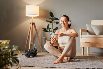 Beautiful smiling woman in headphones looking at smartphone, listening music and relaxing after training wearing beige sportswear enjoying workout.