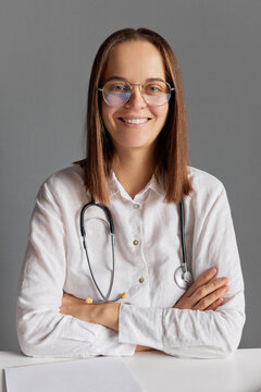 Smiling confident young female doctor in glasses and white medical lab coat looking at camera sitting at office desk against gray wall looking at camera with cheerful face.