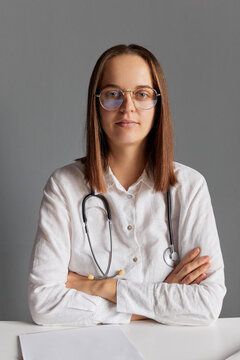 Adult Caucasian doctor in glasses and white medical lab coat looking at camera sitting at office desk against gray wall waiting for her patient keeps hands folded.