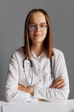 Confident Caucasian female doctor in glasses and white medical lab coat looking at camera sitting at her workplace against gray wall keeps arms crossed.