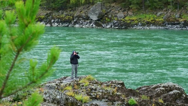 Male photographer taking photos of mountain river landscape while standing on the rocky shore. Traveler man and his camera on the rapid water stream background.