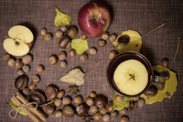 Autumn cozy flatlay composition with apple hot tea, hazelnuts, chestnuts, star anise, cinnamon, fallen leaves on a brown background