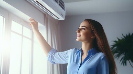 Young attractive woman wearing blue jacket adjusts air conditioner temperature in room at home. - obrazy, fototapety, plakaty
