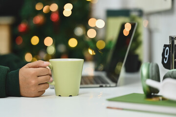 Close up of unrecognizable woman holding cup of hot tea and using laptop near Christmas tree