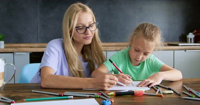 Blonde teacher shows girl pupil how to make picture using color pencil. Schoolgirl with lady teacher sits and draws at desk at home art lesson