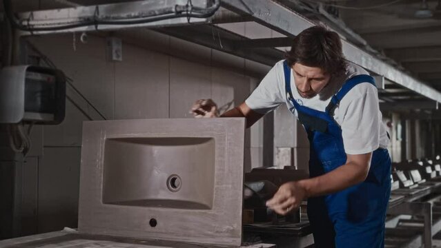 A male worker at a plumbing manufacturing plant processes a sink blank. A professional uses a scraper to remove excess material