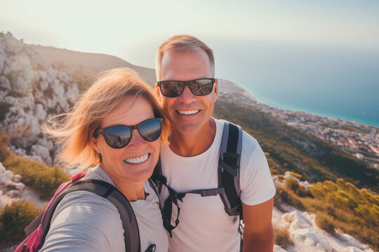 Middle-aged couple with backpacks and sunglasses take a selfie smiling during a route through the mountains with the sea in the background. Couple and sport concept