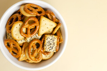 A mix of snacks in a white bowl on a light background. Pretzels with salt, crackers of various shapes and with seeds. Fast food. Harmful snack. Horizontal orientation. Top view. Copy space