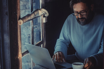 Handsome mature man working with laptop computer in the dark inside a cozy wooden cabin. Concept of...