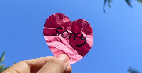 The word sorry written on a pink sticky note in the shape of a heart isolated on a blue sky...