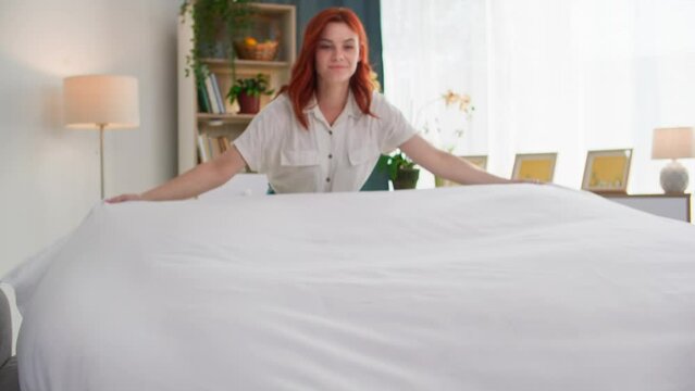 house cleaning, smiling young woman tucking bed linen on bed in cozy bedroom early in morning