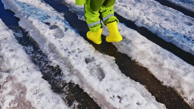 children's feet in yellow rubber boots walk along streams of melting snow on the sidewalk, rear view
