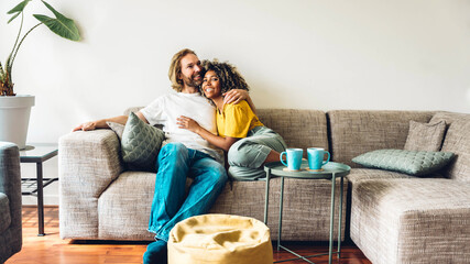 Cheerful multiracial couple sitting on sofa in the living room - Happy family moving in new home -...