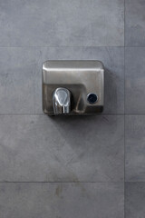 Detail of a hand dryer hanging on a gray tile wall. Front view.