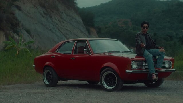 A brutal girl in a brown leather jacket sits on the hood of her vintage red car and smokes