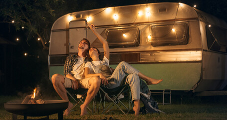 Portrait of a Young Couple Relaxing at a Caravan Camping Area in the Evening, Keeping Warm with Campfire, Looking at the Stars and Dreaming Out Ideas About a Successful Future Together - obrazy, fototapety, plakaty