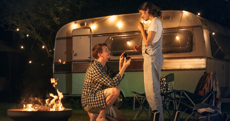 Young Handsome Man Getting on One Knee to Propose to His Beautiful Girlfriend in a Romantic Atmosphere at a Caravan Rest Area with Campfire and Decorative Lights. Fiance Holding an Engagement Ring.