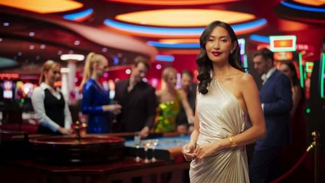 In a Modern Casino Hall: Gorgeous Asian Brunette Woman Posing Confidently, Looking at Camera, Pointing at a Placeholder Space for Advertising or Commercial Materials for a Gambling Industry