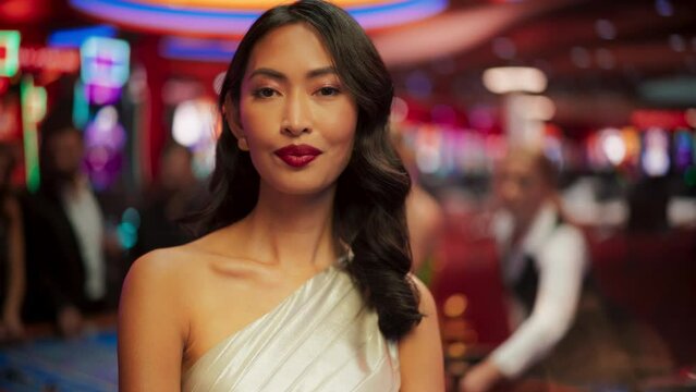 Beautiful Young Asian Woman Standing in a Glamorous Atmosphere of a Casino, Looking at Camera. Anonymous Gamblers Playing in the Background. Close Up Portrait with a Flare Visual Effect
