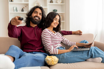 Relaxed millennial couple chilling at home, watching tv, using laptop