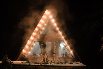 Man hug woman enjoying thermal spa in snowy forest. Couple relax in hot bath outdoors near house with garlands at night. Winter holidays in mountains, hot water treatments concept. Honeymoon vacation