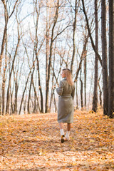 Back view blonde woman in elegant gray coat walks in the sunny autumn season park. Generation z and gen z youth concept.