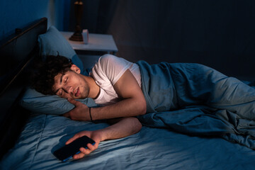 Arabic male lying on comfortable pillow with cellphone in hand and sleeping under blanket at night. Addiction and insomnia